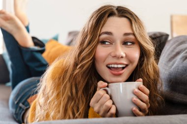 Evdeki kanepede yatarken gülümseyen ve çay içen neşeli güzel kız.
