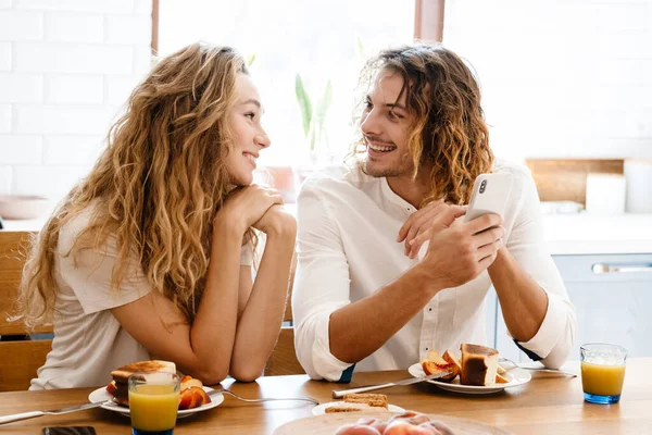 아름다운 부부가 부엌에서 식사를 하면서 미소짓고 핸드폰을 사용하는 — 스톡 사진