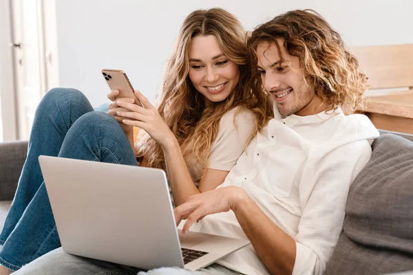 这对年轻貌美的夫妇在家里沙发上休息的时候用手机和笔记本电脑 — 图库照片