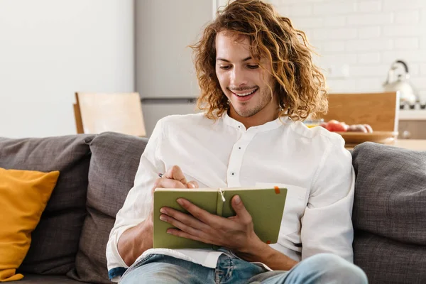 Gelukkig Knappe Man Glimlachen Opschrijven Notities Terwijl Zitten Bank Thuis — Stockfoto