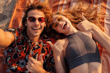 Sahilde uzanmış, kamerayla selfie çeken ve baş parmağını kaldıran neşeli, genç bir çiftin görüntüsü.