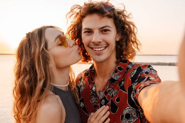 Sahilde fotoğraf çeken ve öpüşen neşeli genç bir çiftin görüntüsü.