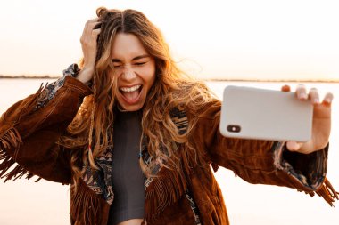 Sahilde yürüyen ve akıllı telefondan selfie çeken heyecanlı genç bir kadının görüntüsü.