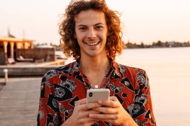 Gün batımında sahilde dururken cep telefonu kullanan gülümseyen genç adam.