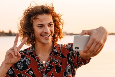 Mutlu genç adam gün batımında sahilde beklerken selfie çekiyor.
