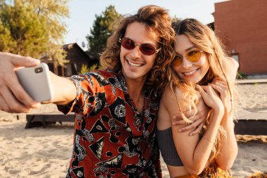 Sahilde cep telefonuyla selfie çeken mutlu, genç bir çiftin fotoğrafı.