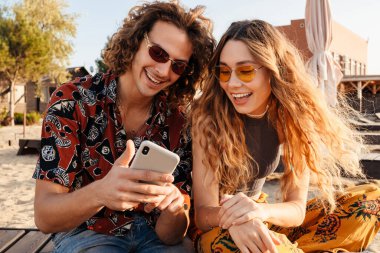 Sahilde cep telefonu kullanan mutlu, genç bir çiftin görüntüsü.