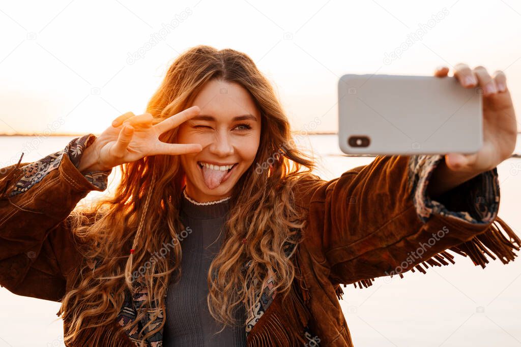 Photo of a positive young woman walking outdoors at the beach and taking a selfie by smartphone and showing peace gesture