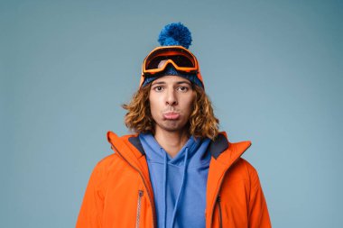 Mavi arka planda yalnız duran, kışlık ceket giyen üzgün bir delikanlıya yaklaş.