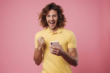 Portrait of an excited young man using mobile phone isolated over pink background, celebrating
