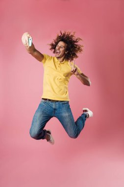 Pembe arka planda izole edilmiş cep telefonundan selfie çekerken barış işareti gösteren heyecanlı yakışıklı adam.