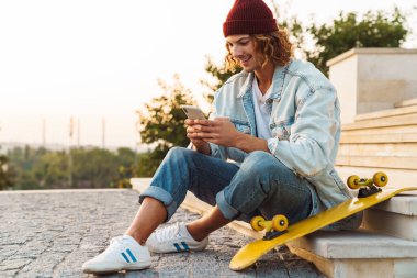 Neşeli, pozitif, kıvırcık bir adamın açık havada oturup cep telefonu kullanması.