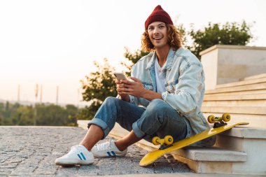 Neşeli, pozitif, kıvırcık bir adamın açık havada oturup cep telefonu kullanması.