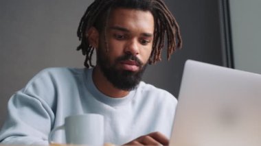 Memnuniyet duyan Afro-Amerikan bir adam dizüstü bilgisayarında daktilo yazıyor. Evdeki masada oturuyor.