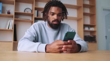 Odaklanmış, Afro-Amerikan bir adam dairesinde oturan akıllı telefonunu kullanıyor.