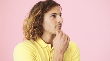 Düşünen kıvırcık genç bir adam bir fikir ileri sürerken parmağını kaldırıyor ve stüdyoda pembe bir arka planda tek başına duran kameraya bakıyor.