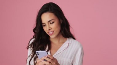 Güzel, genç bir kadın akıllı telefonunu kullanıyor ve stüdyoda pembe bir arka planda tek başına ayakta duruyor.