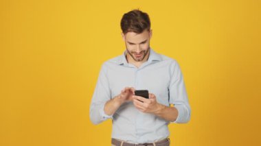 Gülümseyen genç bir adam akıllı telefonunu stüdyodaki sarı arka planda duruyor.