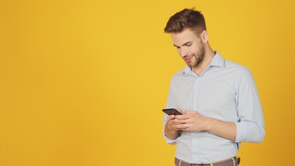 一个快乐的年轻人正在用他的智能手机站在独立于工作室黄色背景之外的一块空地上 — 图库视频影像