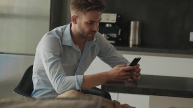 Ciddi bir genç işadamı akıllı telefonunu evde mutfakta otururken kullanıyor.