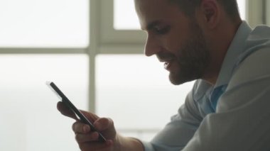 Sakin genç bir iş adamının yakın çekim görüntüsü akıllı telefonuyla evde mutfakta oturuyor.