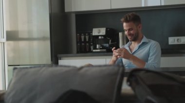 Memnun genç bir işadamı akıllı telefonunu evde mutfakta otururken kullanıyor.