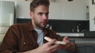 Pozitif bir genç adam evde oturan biriyle konuşurken pizza yiyor.