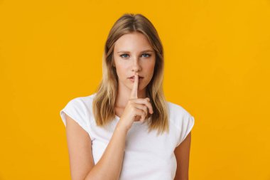 Ciddi, güzel bir kız, sarı arka planda izole edilmiş kameraya sessiz jest yapıyor.