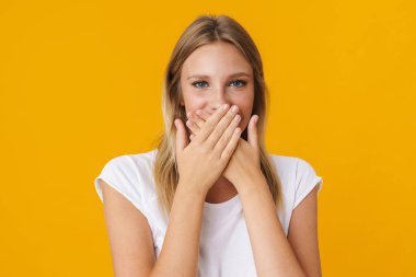 Neşeli güzel kız poz veriyor ve ağzını kapatıyor. Sarı arka planda izole edilmiş.