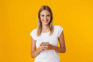 Sarı arka planda izole edilmiş gülümseyen ve cep telefonu kullanan neşeli sarışın kız.