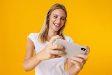 Sarı arka planda izole edilmiş cep telefonuyla oyun oynayan neşeli sarışın kız.