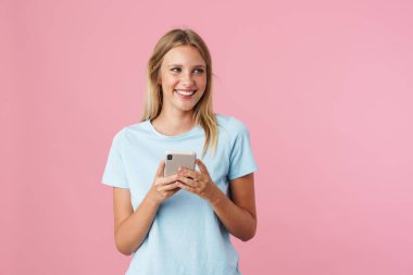 Gülümseyen genç bir kadın pembe arka planda tek başına dururken cep telefonu kullanıyor.