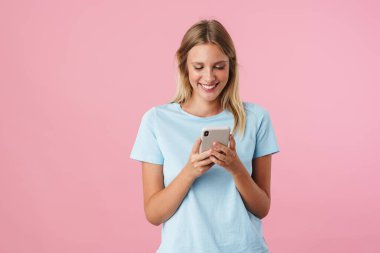 Gülümseyen genç bir kadın pembe arka planda tek başına dururken cep telefonu kullanıyor.