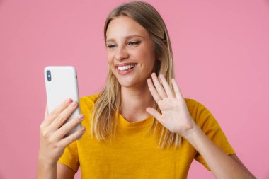 Neşeli kız pembe arka planda izole edilmiş cep telefonuyla selfie çekerken el sallıyor.