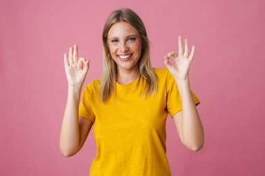 Neşeli güzel kız gülümsüyor ve pembe arka planda iyi bir işaret gösteriyor.