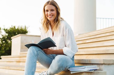 Güzel mutlu öğrenci kız dışarıda merdivenlerde otururken egzersiz defterine notlar yazıyor.