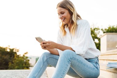 Dışarıda merdivenlerde otururken gülümseyen ve cep telefonu kullanan mutlu bir kadın.