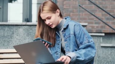 Güzel bir genç kadın, cep telefonu ve dizüstü bilgisayarını kullanıyor. Sokakta bankta oturuyor.