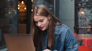 Düşünen genç bir kadın şehir kafesinde otururken dizüstü bilgisayarını kullanıyor.