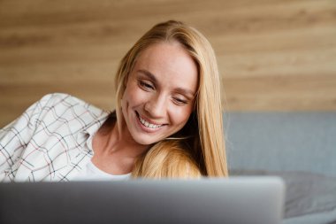 Güzel sarışın gülümseyen kadın evde yatarken dizüstü bilgisayar kullanıyor.