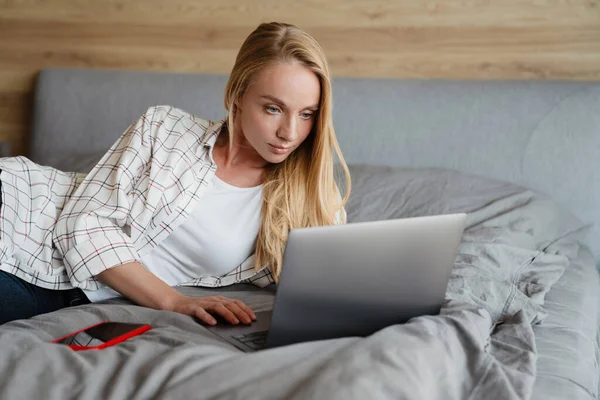 Hermosa Mujer Rubia Enfocada Utilizando Ordenador Portátil Mientras Está Acostado — Foto de Stock