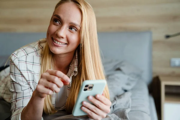 Szőke Mosolygós Fülhallgatóban Mobiltelefont Használ Miközben Otthon Fekszik Ágyon — Stock Fotó