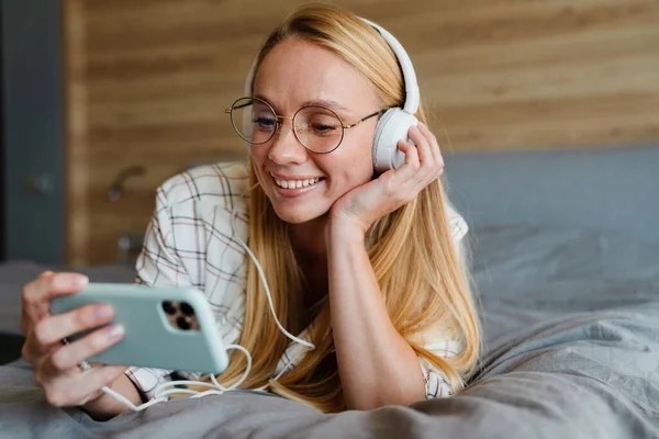 Mujer Rubia Sonriente Escuchando Música Con Auriculares Teléfono Móvil Cama —  Fotos de Stock