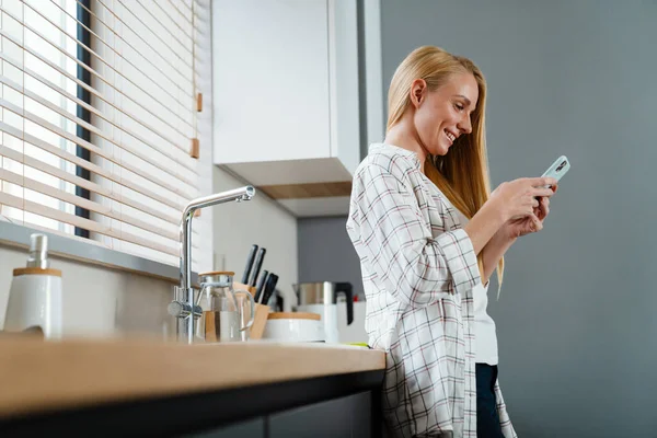 Blond Lykkelig Kvinne Som Bruker Mobiltelefon Smiler Kjøkkenet Hjemme – stockfoto