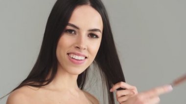 Güzel, yarı çıplak, uzun siyah saçlı bir kadın, saçlarını tarıyor. Stüdyonun gri arka planında tek başına duruyor.