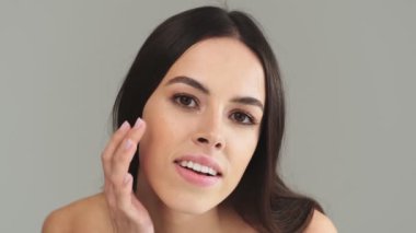 Pozitif yarı çıplak, uzun siyah saçlı bir kadın kameraya bakıyor. Stüdyonun gri arka planında tek başına duran aynaya bakıyor.