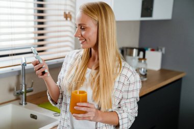 Sarışın mutlu kadın cep telefonu kullanıyor ve evde meyve suyu içiyor.