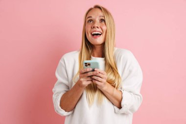 Heyecanlı güzel kız gülümsüyor ve cep telefonunu pembe arka planda kullanıyor.