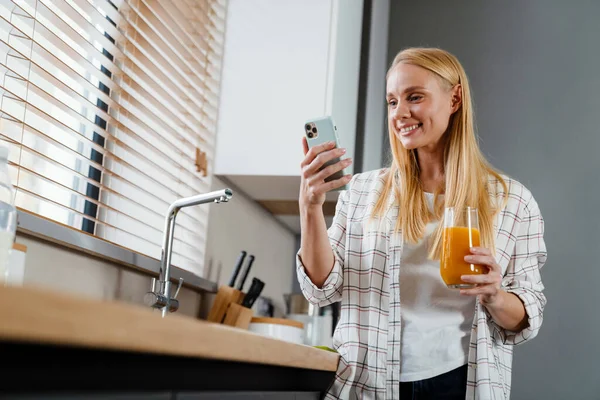 Счастливая Блондинка Пользуется Мобильным Телефоном Пьет Сок Домашней Кухне — стоковое фото