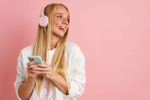 幸せな美しいです女の子リスニング音楽とともにヘッドフォンと携帯電話隔離上のピンクの背景 — ストック写真